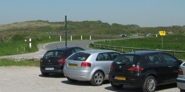 a corner of a road among dunes with cars in a car park in the foreground
